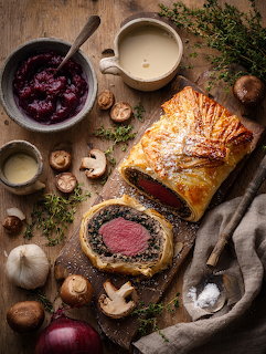 Sliced beef Wellington with golden pastry on a cutting board surrounded by mushrooms, herbs, and cranberry sauce