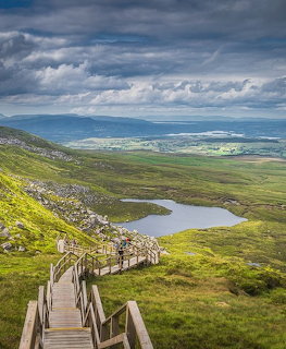 Wooden staircase on Cuilcagh Mountain with lake and green valley below under dramatic clouds