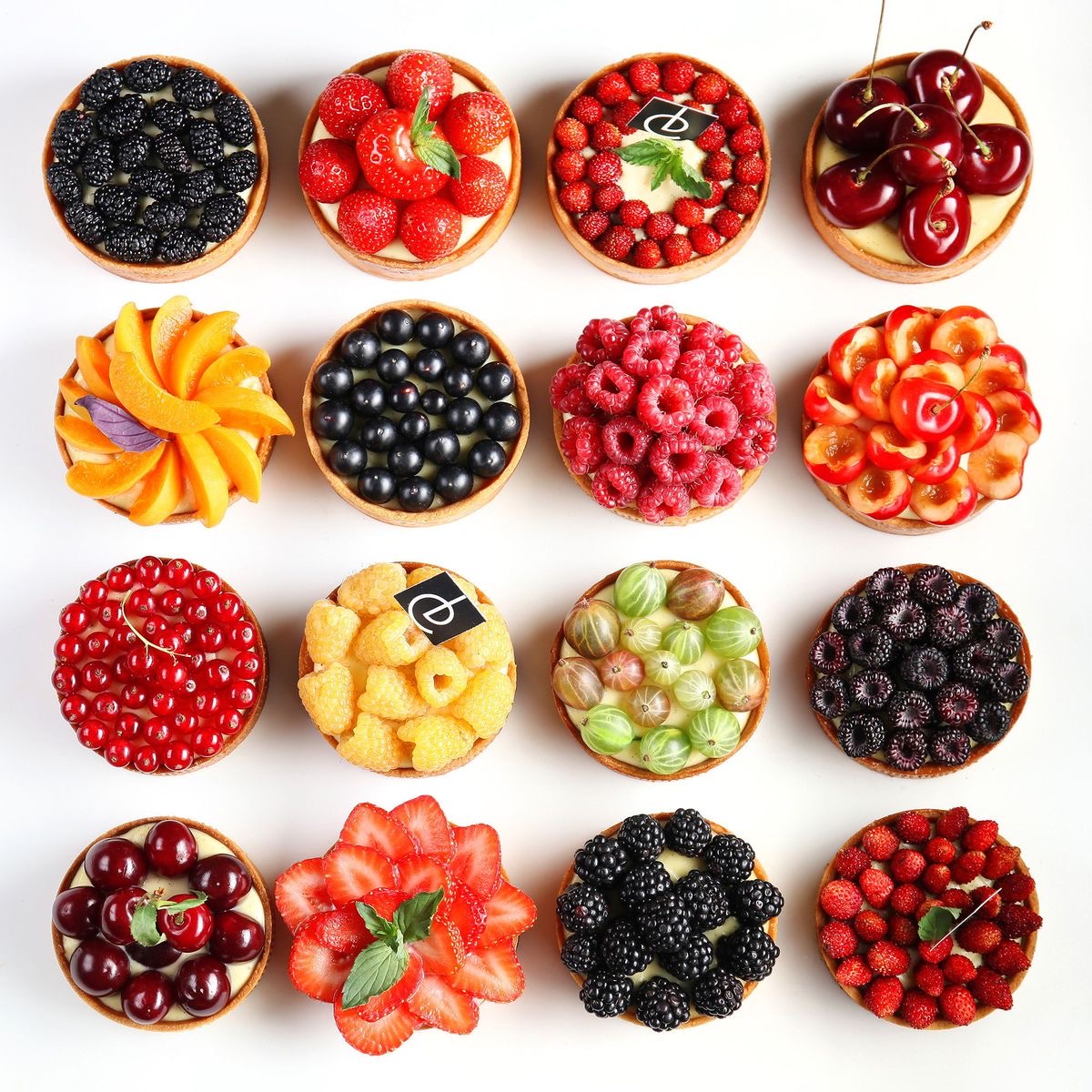 Assorted fruit tarts with various berries arranged in rows on white background