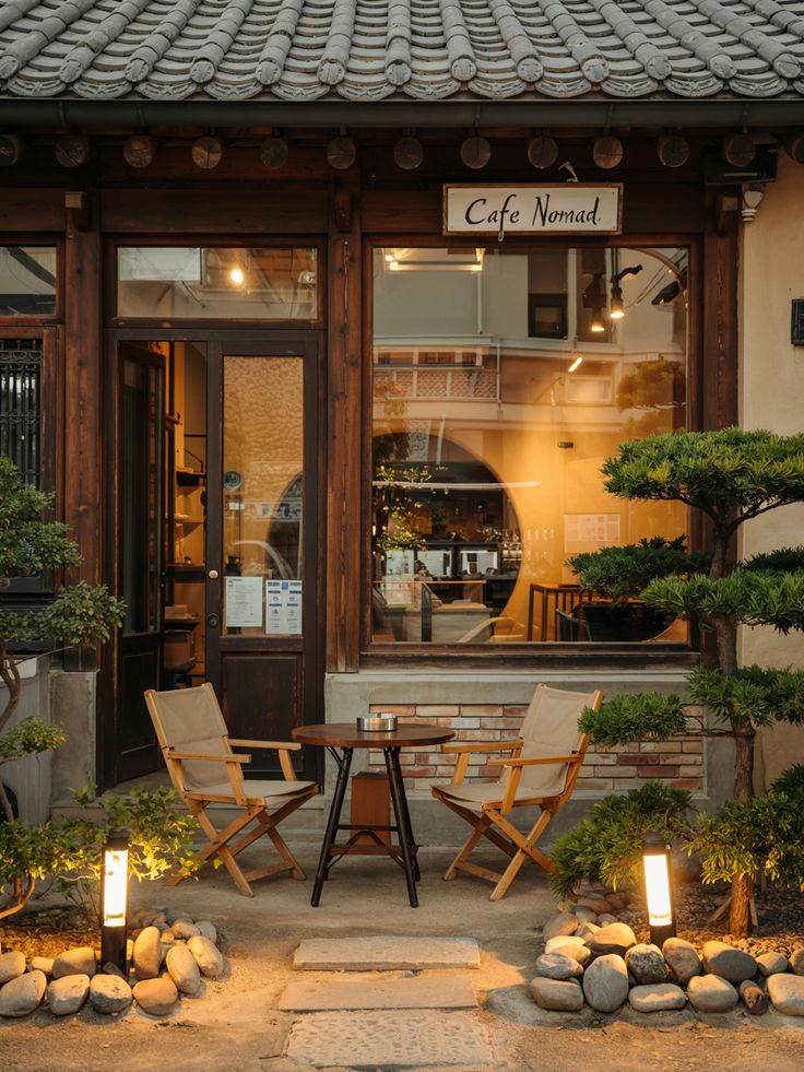 Cafe Nomad storefront with bonsai trees, wooden chairs, stone path, and warm glowing lights at dusk