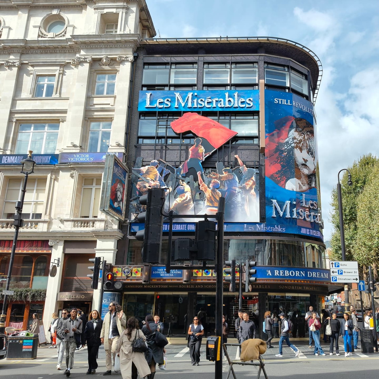 City street corner with a theater building displaying large Les Misérables posters and people walking outside