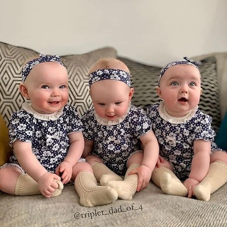 Three smiling baby triplets wearing matching black floral outfits sitting on a couch
