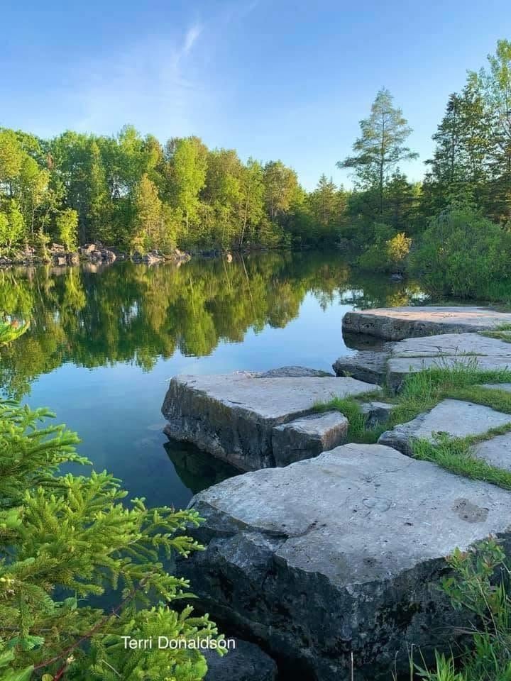 Flat rocks beside a calm lake with green trees reflected in the water under a blue sky