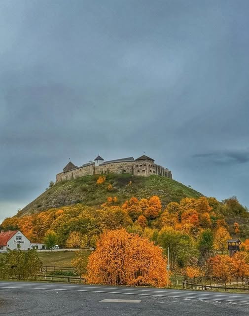 Medieval stone castle on a forested hilltop with orange and yellow autumn trees under a cloudy sky