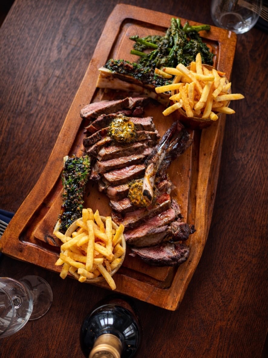 Sliced grilled steak with herb butter on a wooden cutting board with fries and broccolini
