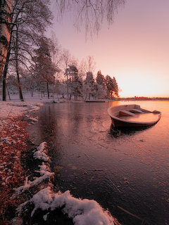 Snow-covered lake shore at sunset with a boat and distant castle island under colorful sky