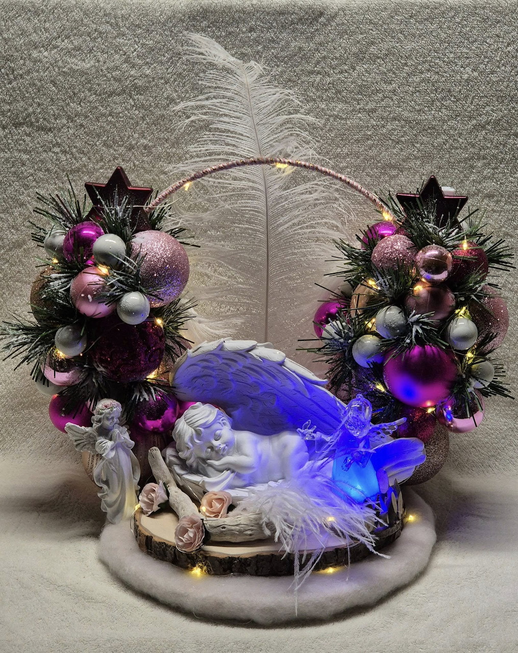 Christmas decoration with sleeping angel, blue wings, pink and purple ornaments, fairy lights, and white feather