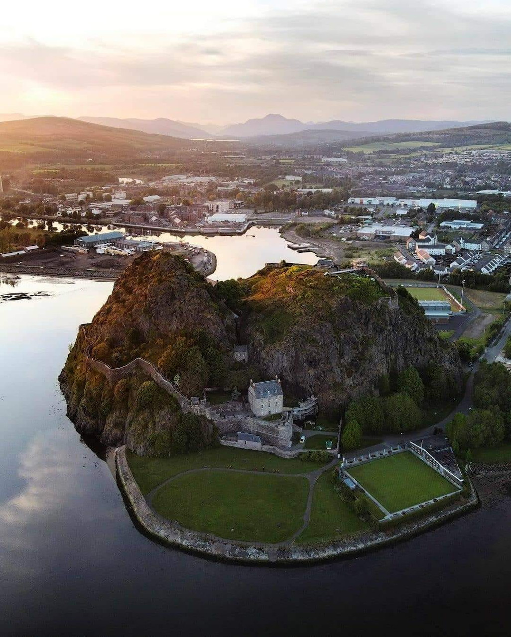 Aerial view of Dumbarton Castle on a rocky outcrop surrounded by water with Scottish landscape and sunset sky