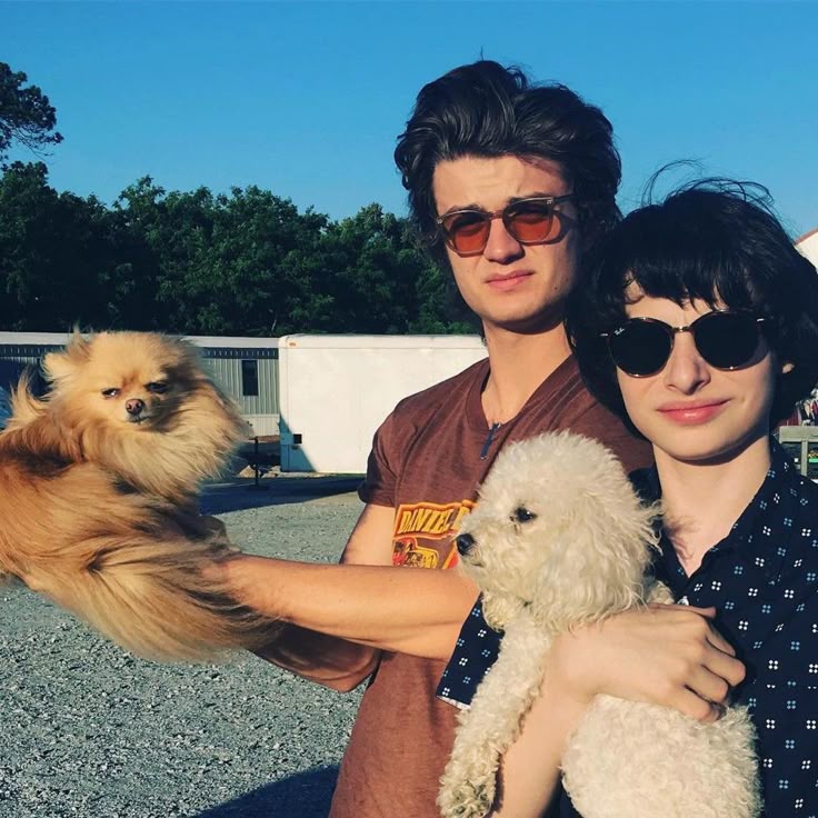 Joe Keery and Finn Wolfhard wearing sunglasses and holding two fluffy dogs outdoors