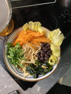 Bowl of ramen with noodles, black beans, carrots, lettuce, cilantro, and seaweed on a dark surface