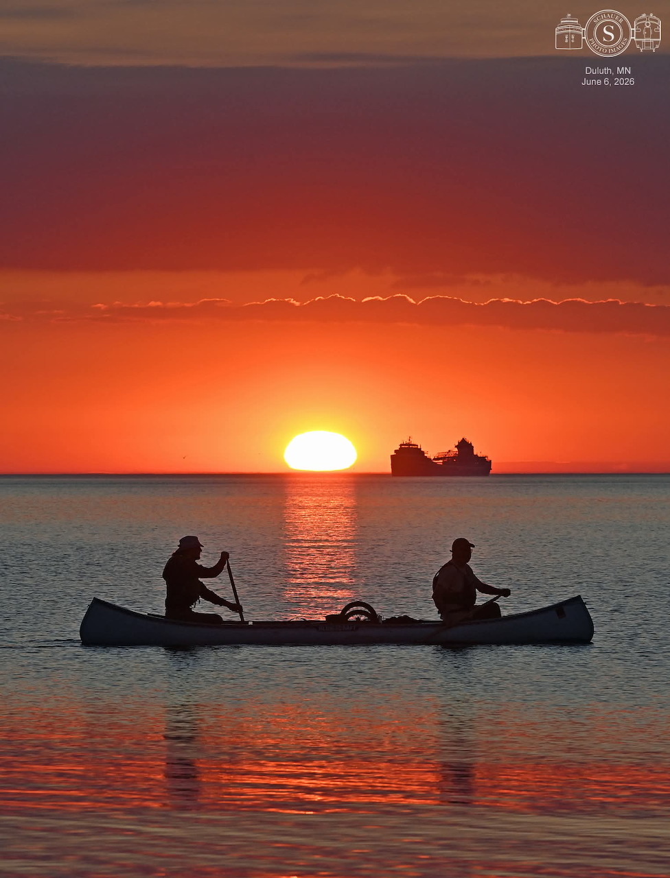 Two silhouetted canoeists on calm water at sunset with ships on the horizon