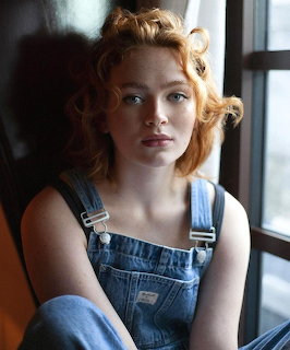 Young woman with red wavy hair in denim overalls sitting by window