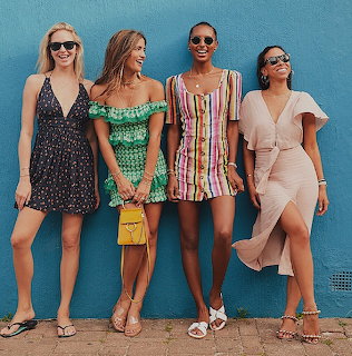 Four women in sunglasses wearing colorful summer dresses standing against a blue wall