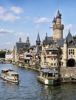 Castle-style riverside building with towers beside a river and boats on the water