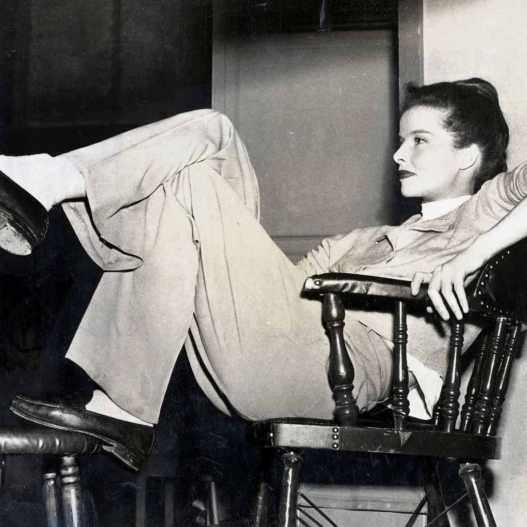 Black and white photo of Katharine Hepburn reclining on wooden chair in casual clothing