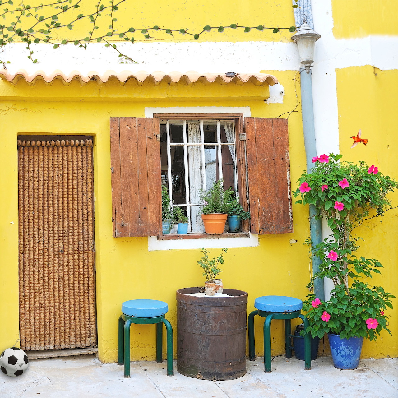 Vibrant yellow house exterior with wooden shutters, bamboo door, window with plants, and blue stools around a barrel table