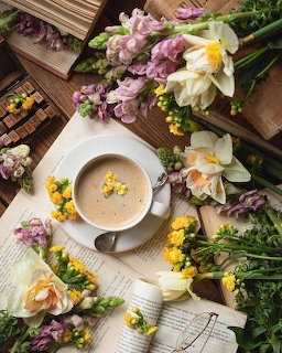 Cup of coffee on saucer surrounded by daffodils, pink flowers, and open books on a wooden table