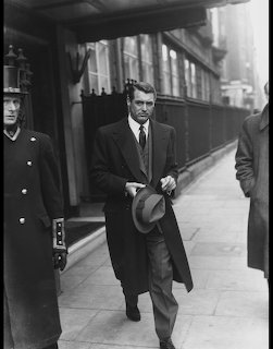 Black-and-white photo of Cary Grant walking on a city sidewalk in an overcoat and tie, holding a hat.