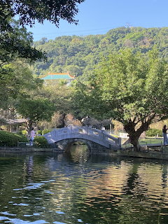 Stone arch bridge over a reflective pond surrounded by green trees in a traditional Chinese garden