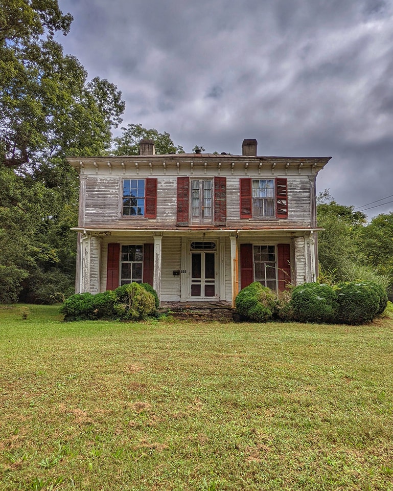 Abandoned two-story white farmhouse with red shutters and front porch on a green lawn under cloudy skies