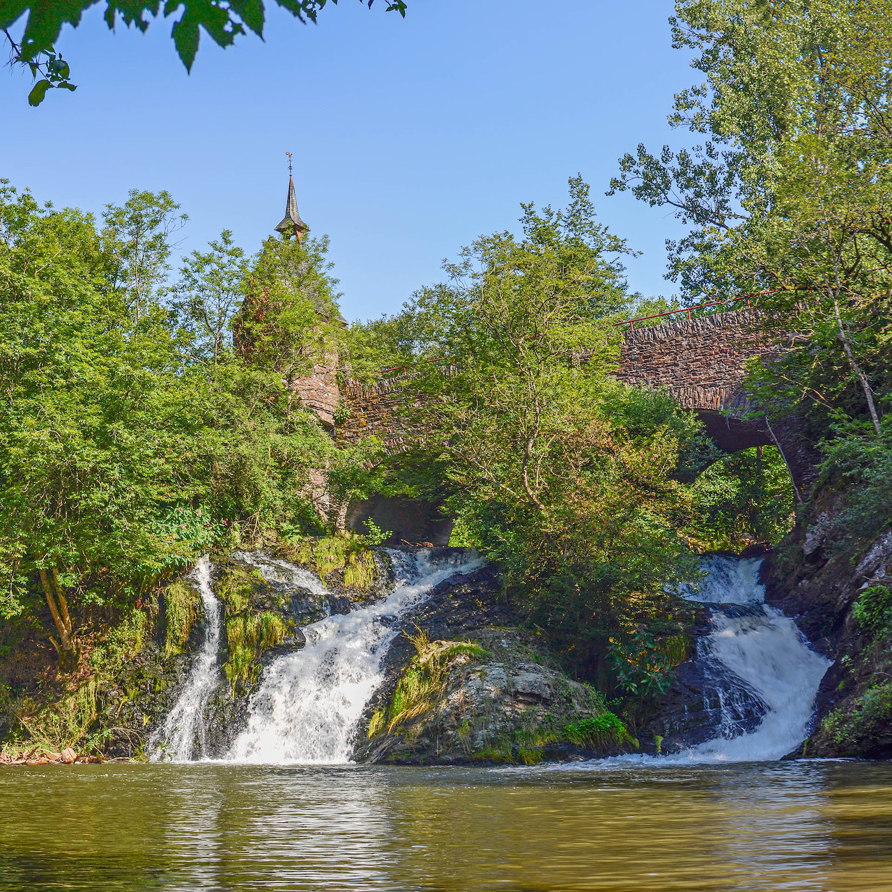 Small waterfall flowing into a pond among green trees, with a roof and spire partly visible in back