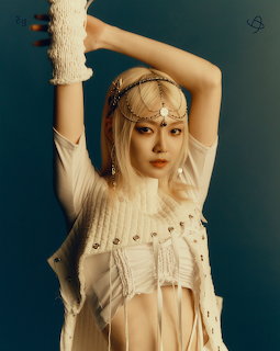 Young woman with blonde hair wearing white knit top and ornate chain headpiece posing with arms raised