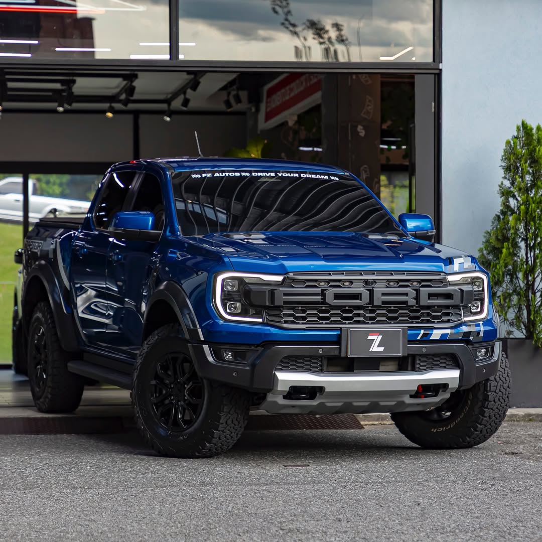 Blue Ford Raptor pickup truck in showroom with black wheels and chrome grille