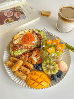 Colorful breakfast plate with fried egg on avocado toast, shrimp, mango, fruit cup, and a latte