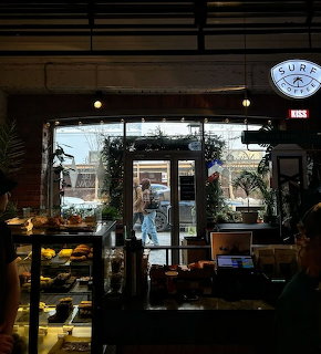 Interior of a coffee shop with pastry display case, plants, string lights, and a large window to the street
