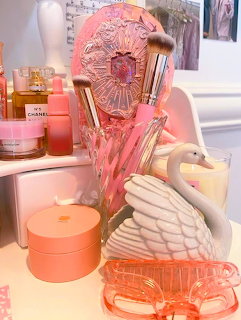Pink vanity table with Chanel perfume, makeup brushes, white swan figurine, candles, and coral accessories