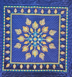Blue quilt with golden leaf and tulip appliqués arranged in a radial star flower pattern with decorative border
