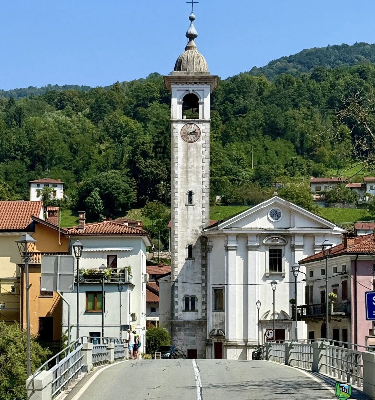 Stone bridge with tall bell tower and white church facade in Slovenian village surrounded by green hills