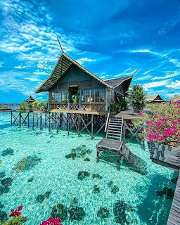 Wooden overwater bungalow on stilts above clear turquoise water with pink flowers and blue sky