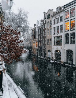 Snow-covered Amsterdam canal with traditional Dutch buildings and falling snowflakes in winter