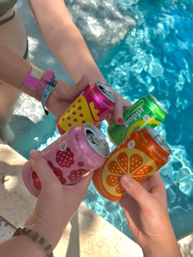 Four hands holding colorful fruit soda cans above bright blue pool water