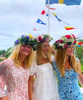Three blonde women in white dresses with vibrant flower crowns at outdoor Midsommar festival