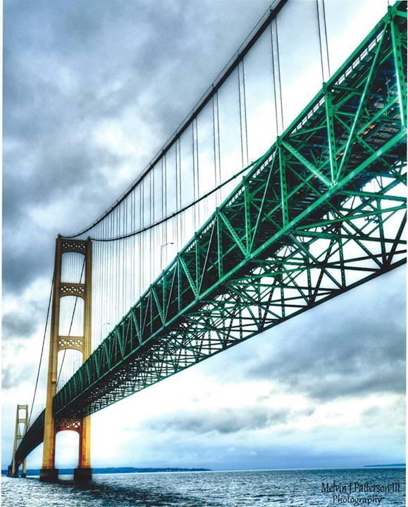 Green suspension bridge with tall towers spanning over blue water beneath cloudy sky