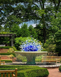 Large ornate blue and yellow glass sculpture in sunlit garden with green hedges and wooden benches