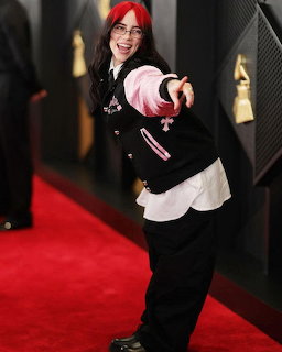 Billie Eilish with red and pink hair posing on a red carpet in a black jacket and white shirt