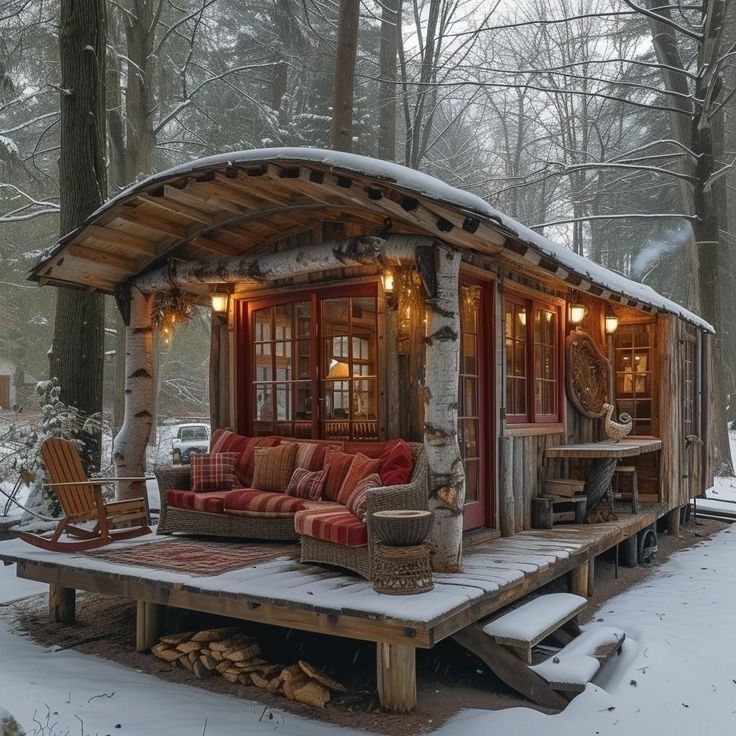 Rustic cabin with curved metal roof on wooden deck in snowy forest with warm interior lighting