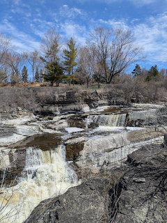 Rocky waterfall flowing over layered stone ledges with trees and blue sky in early spring