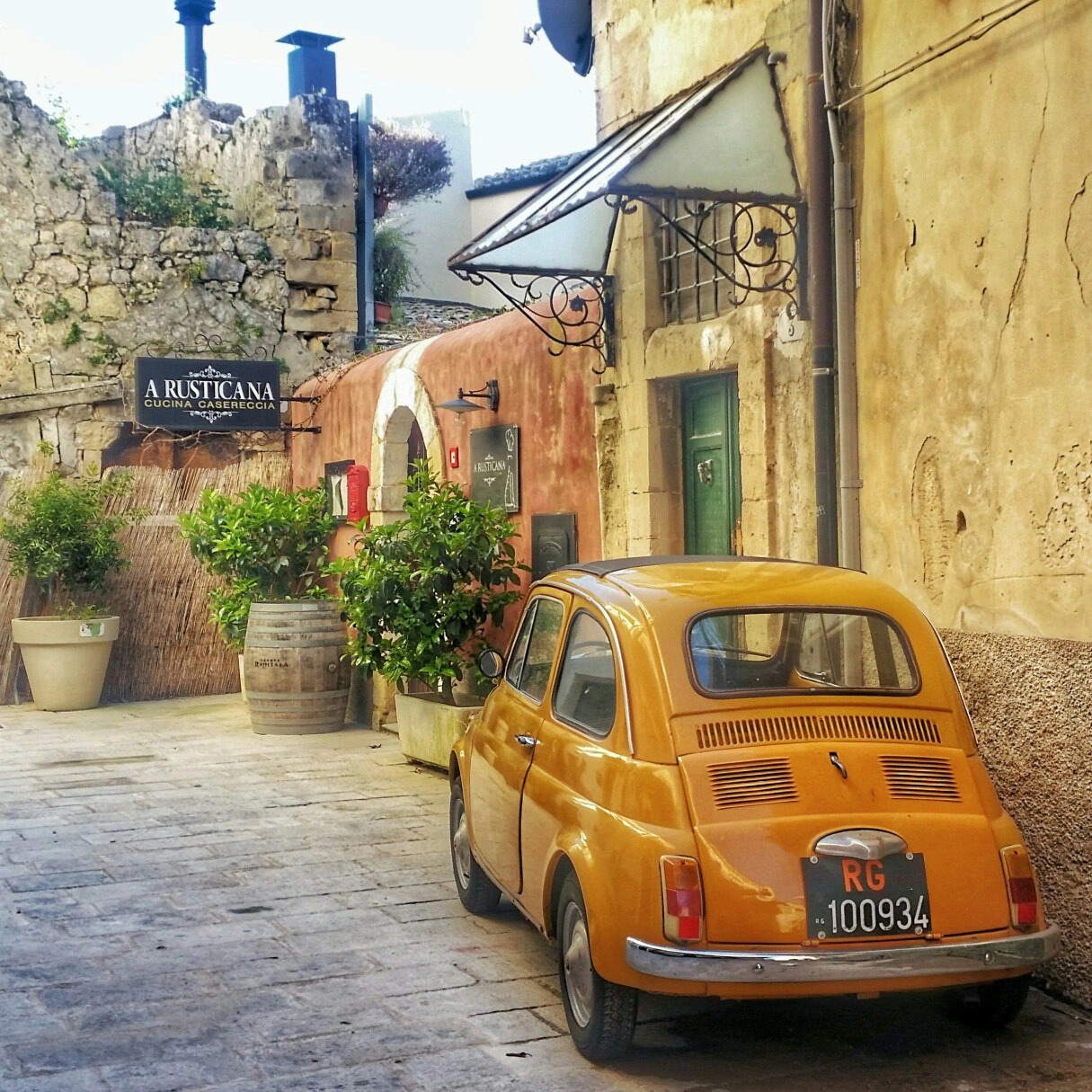 Vintage yellow Fiat 500 parked on a narrow European street with stone buildings and flower pots