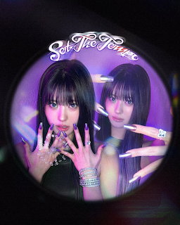 Two women with long dark hair posing inside a circular lens frame with purple neon lighting