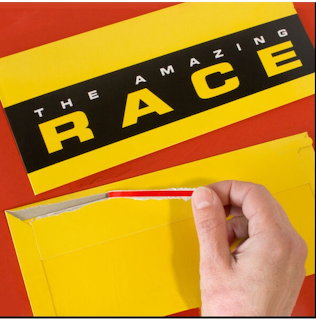 Hand opening a yellow envelope with a The Amazing Race card on a red background