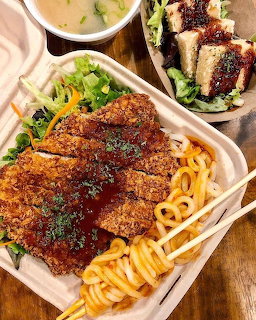Takeout bento box with breaded katsu cutlet on spaghetti, soup bowl, and salad on wooden table