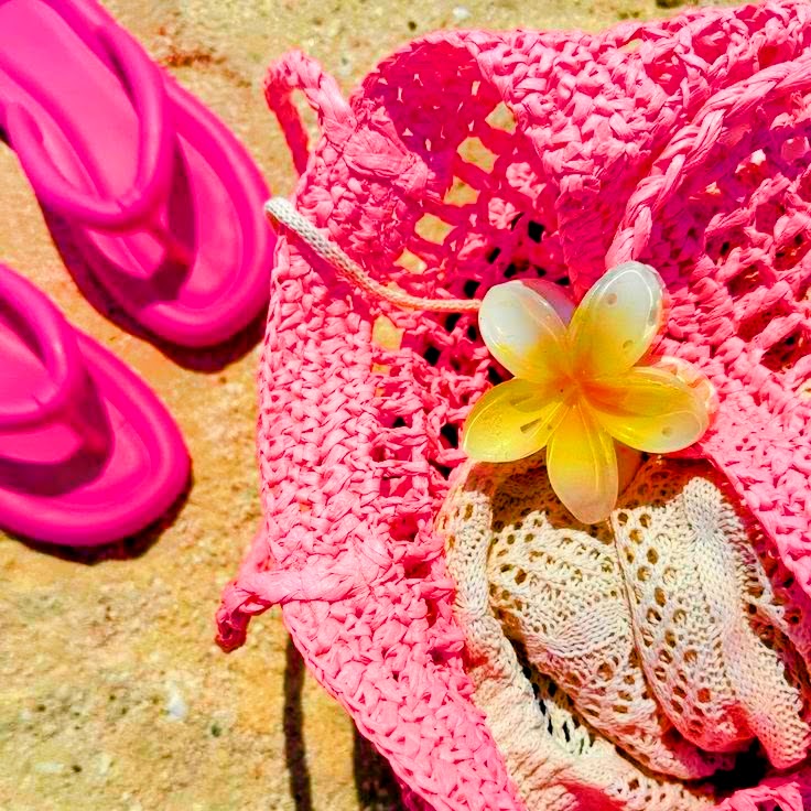 Pink flip-flops, beach bag, and yellow flower on sand with tropical accessories