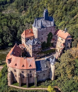 Aerial view of a medieval stone castle with orange roofs surrounded by green forest
