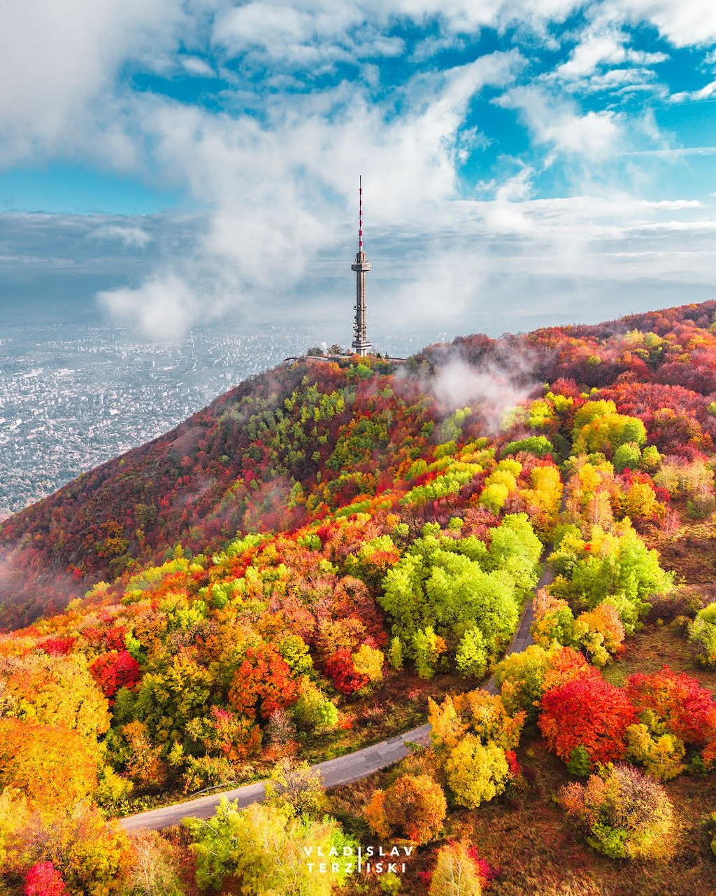 Aerial view of a TV tower atop a hill covered in red, orange, and green autumn trees with a city in the background.