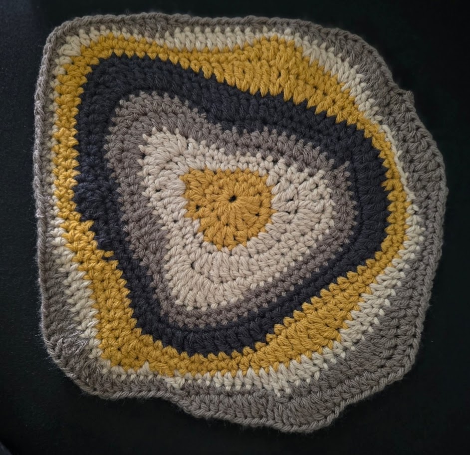 Crocheted granny square with mustard, gray, and cream concentric rounded layers on dark background