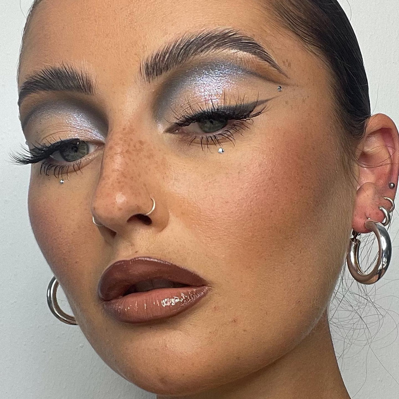 Close-up of woman's face with dewy makeup, winged eyeliner, and glossy lips wearing silver hoop earrings